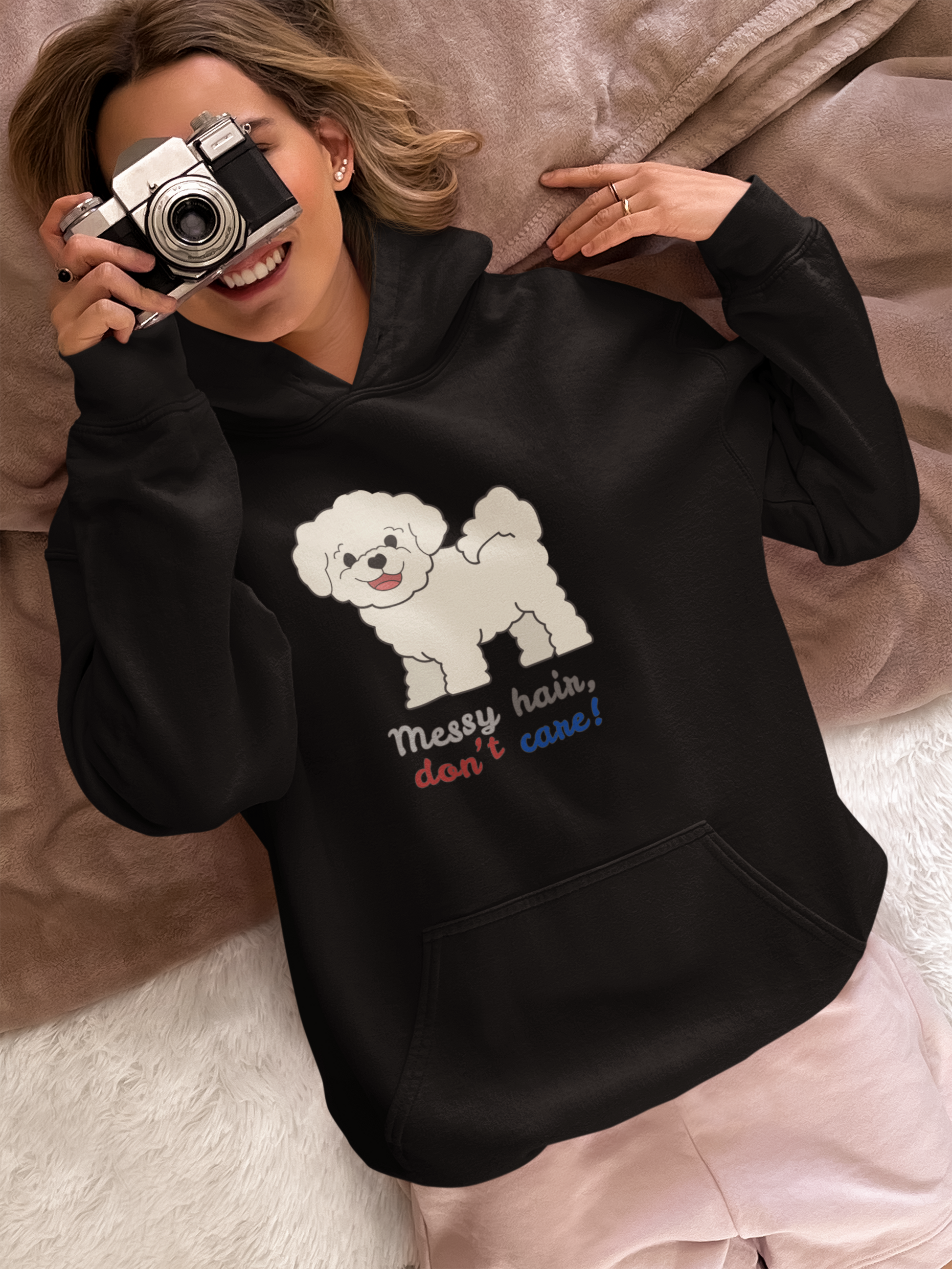 Woman lying in bed wearing bichon dog hoodie with messy hair don’t care slogan while pointing at the ceiling with a vintage camera