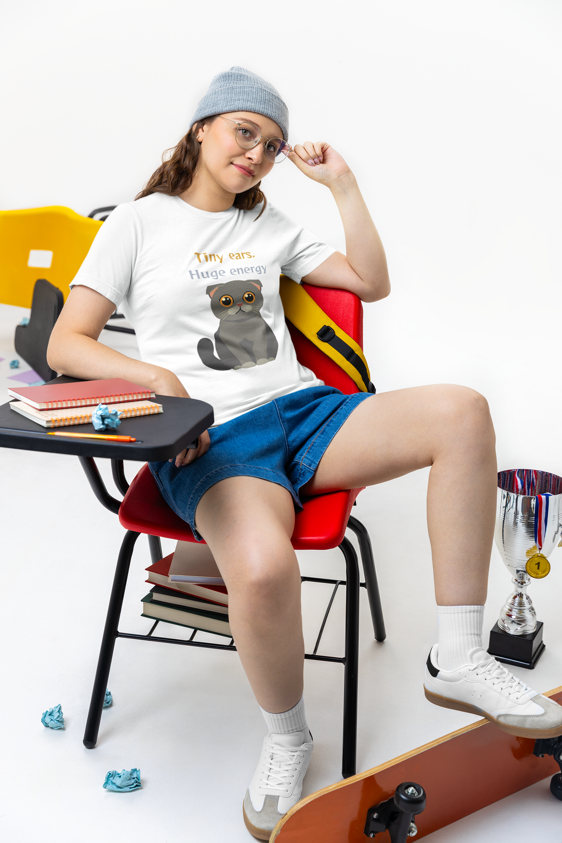 Young woman student posing in a school classroom wearing white t-shirt with Scottish Fold cat graphic and tiny ears huge energy slogan