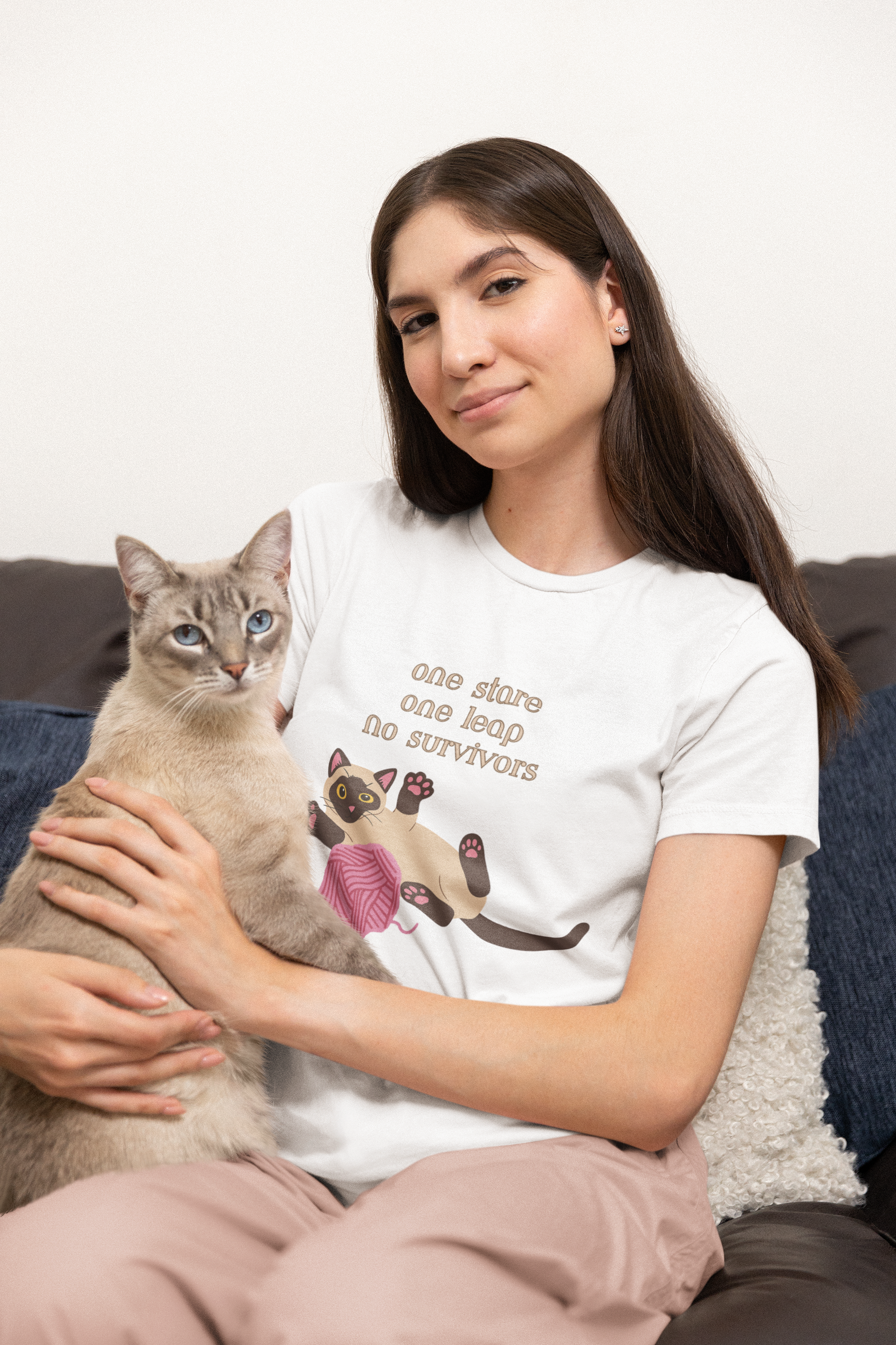 Happy woman holding a cute cat while wearing white t-shirt with Siamese cat graphic and one stare one leap no survivors slogan